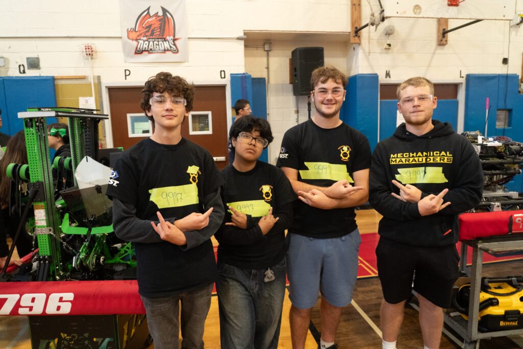 Four Team 271 students posing with robots in a school gym